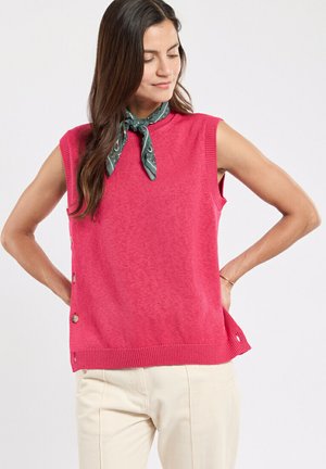 Femme portant un gilet sans manches rose avec des boutons latéraux, un foulard vert à motifs autour du cou, et un pantalon beige clair, debout les mains sur les hanches.