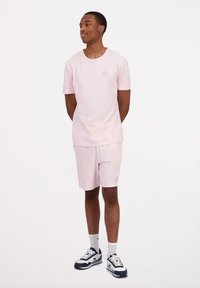 Completo in cotone di colore rosa chiaro composto da t-shirt e pantaloni corti. La t-shirt presenta un piccolo logo sul petto. Il modello indossa calze bianche e scarpe da ginnastica blu navy.