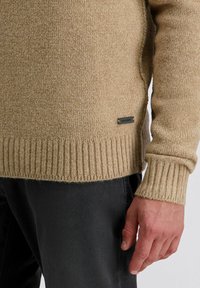 Beige gestrickter Pullover mit gerippten Bündchen und Saum, versehen mit einem kleinen schwarzen Etikett an der Seite. Weicher Stoff und lässige Passform.