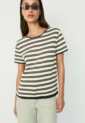 Femme aux longs cheveux foncés portant des lunettes de soleil rectangulaires, un t-shirt blanc à manches courtes avec des rayures horizontales noires et vert olive, et un pantalon clair.