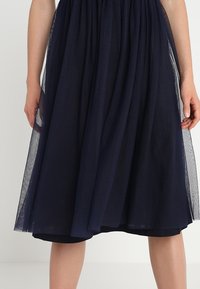 Jupe en tulle marine avec un design superposé, offrant une texture douce et une ceinture élastique pour un ajustement confortable. Sa longueur descend en dessous du genou.