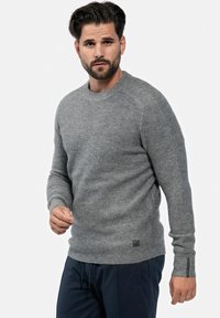 Grauer gestrickter Pullover mit Rundhalsausschnitt, gerippten Bündchen und Saum sowie einem kleinen Logotag am unteren Rand. Getragen mit dunkelblauen Hosen.