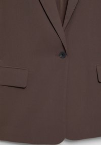Veste de costume marron avec un unique bouton noir et deux poches à rabat devant, montrant la partie inférieure du torse.