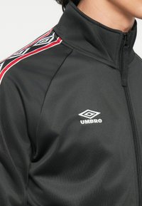 Črna jakna z visokim ovratnikom, ki ima rdeče in modre diagonalne črte ter vezeno belo logo Umbro na prsih.