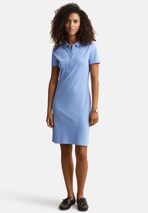 Frau mit lockigem Haar, die ein knieumspieltes hellblaues Polokleid und schwarze Schuhe trägt, stehend vor einem schlichten weißen Hintergrund.