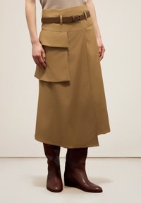 Jupe midi kaki avec une ceinture, dotée d'une grande poche avant et d'un ourlet dessiné. Associée à des bottes marron foncé, montantes jusqu'au genou. Texture lisse.