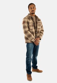 Chaqueta a cuadros marrón con botones y dos bolsillos en el pecho, llevada sobre una camisa beige, combinada con jeans azules y botas marrones de cordones. Fondo blanco.