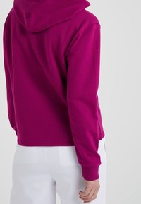 Sweat à capuche fuchsia à manches longues, coupe décontractée, poignets côtelés et texture lisse, porté avec un pantalon blanc.