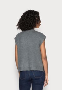 Harmaa neulottu hihaton villapaita, jossa resorikaulus ja -helma, korostaen istuvaa muotoa ja pehmeää koostumusta. Yhdistetty tummiin farkkuihin.