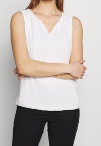 Blouse blanche à manches courtes avec un décolleté en V, fabriquée dans un tissu lisse. Présente un drapé subtil au niveau du décolleté et une coupe décontractée.