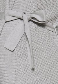 Närbild av ett ljusgrått stickat tyg med ett ribbat mönster och en knuten rosett gjord av samma material.