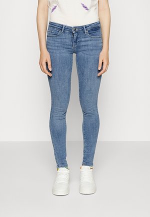 Personne portant un jean skinny bleu clair, des baskets blanches et un haut blanc avec une broderie florale partielle, debout devant un fond uni.