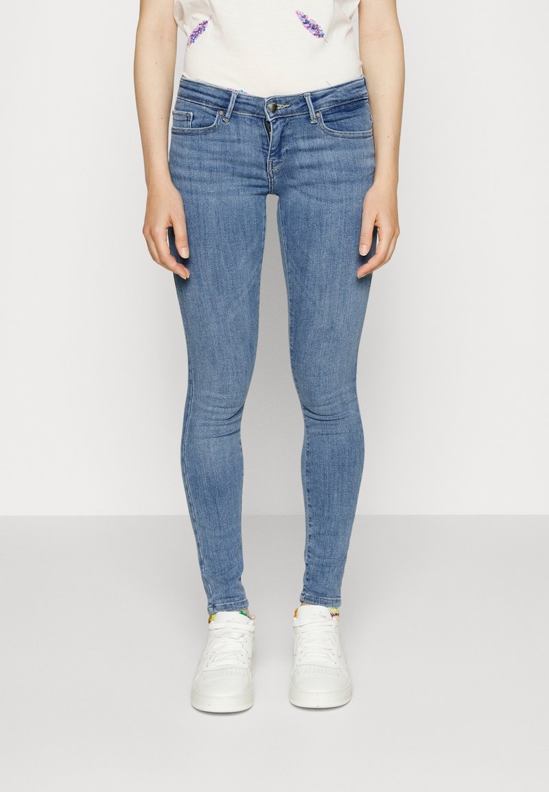 Personne portant un jean skinny bleu clair, des baskets blanches et un haut blanc avec une broderie florale partielle, debout devant un fond uni.
