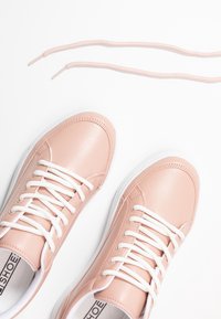 Paire de baskets rose clair avec des lacets blancs sur fond blanc, un lacet partiellement défait et étendu au-dessus des chaussures.
