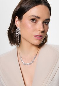 Srebrne kolczyki w kształcie liści oraz pasujący naszyjnik, na którym widać misterną detalizację i polerowane wykończenie, noszone przez modelkę na neutralnym tle.