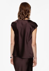 Blusa in raso marrone con maniche corte a cappuccio e scollatura tonda. Il tessuto ha una consistenza liscia e una leggera lucentezza, caratterizzato da un design fluido.