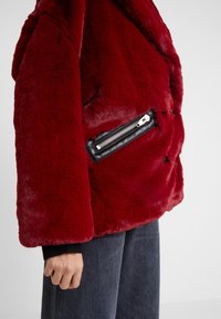 Röd fuskpälsjacka med en svart läderficka med dragkedja. Har en bred krage och en mjuk, plyschliknande textur. Ärmarna är löst sittande.