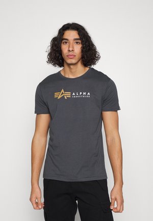 Tricou din bumbac gri, cu un logo imprimat aurit care spune "ALPHA INDUSTRIES" și un design cu stea pe față. Guler clasic tip crew.