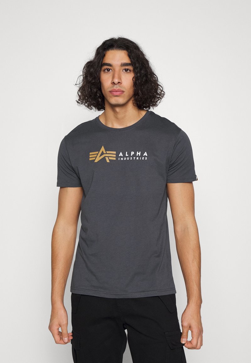 Šedé bavlněné tričko s vytištěným zlatým logem s nápisem "ALPHA INDUSTRIES" a designem hvězdy na přední straně. Klasický kulatý výstřih.