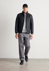 Young man walking indoors wearing black cap, black quilted jacket, white shirt, gray pants, and black sneakers.
