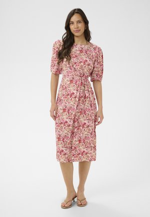 Kobieta stojąca w różowej sukience midi w kwiaty z bufkami i przewiązanym w talii paskiem, w beżowych sandałach na obcasie na białym tle.
