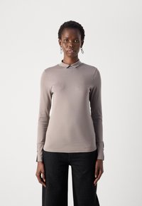 Femme aux cheveux courts portant un polo à manches longues taupe et un pantalon large noir à pois, se tenant devant un fond clair.
