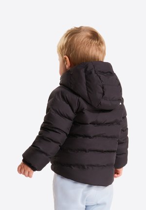 Niño pequeño con el cabello rubio corto, vestido con una chaqueta acolchada negra con capucha y pantalones de color claro, de espaldas.