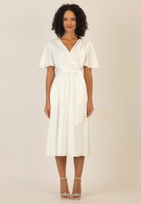 Weißes Wickelkleid mit kurzen Schmetterlingsärmeln, gebundenem Taillenband und fließendem Rock. Aus glattem Stoff mit knielangem Saum. Silberne Riemchensandaletten.