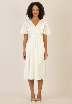 White wrap dress with short flutter sleeves, tie waist, and flowing skirt. Features smooth fabric and a knee-length hem. Silver strappy heels.
