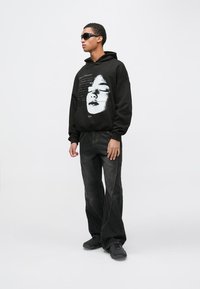 Mladi moški v črni kapucarji z belim odtisom obraza, širokih temnih kavbojkah, črnih supergah in črnih sončnih očalih, stoji ob preprosti ozadju.