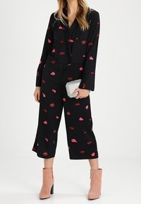 Woman wears black cropped jumpsuit with red and pink lip prints, holding silver clutch, paired with light pink suede ankle boots.