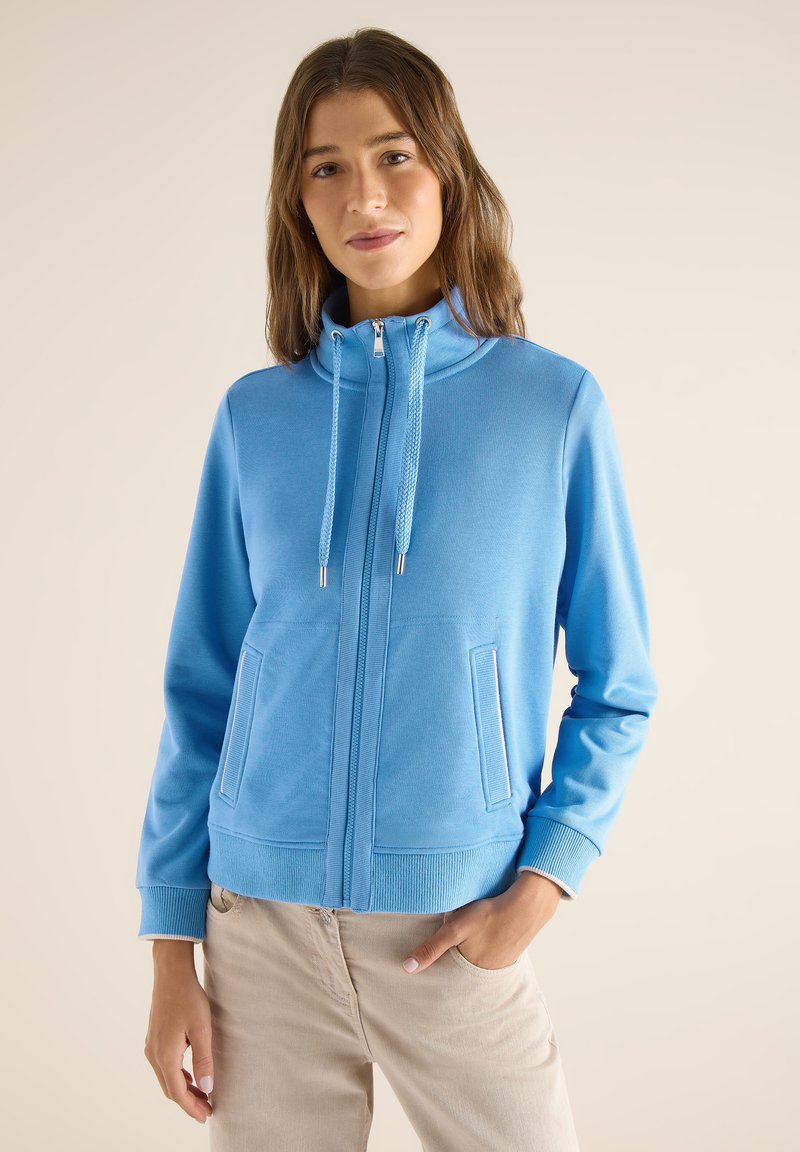Jeune femme portant un sweat-shirt zippé bleu clair avec cordons et un pantalon beige, debout avec une main dans la poche sur un fond uni.