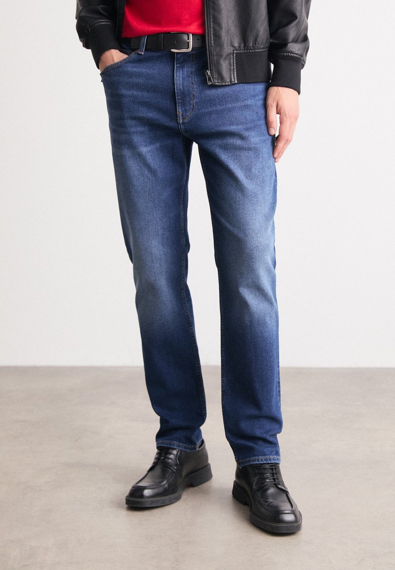 Modri kavbojski ovan s ravnim krojem, ki vključuje klasično zasnovo s petimi žepi in rahlim zbledelostjo, v kombinaciji s črnimi usnjenimi čevlji.