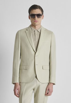 Man in een beige pak en bijpassend poloshirt met donkere zonnebril, staand tegen een effen lichtgrijze achtergrond.