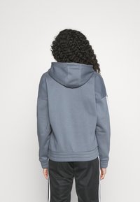 Sweat à capuche gris avec une grande capuche, un ourlet et des poignets côtelés. Présente une texture lisse et un design minimaliste. Associé à un pantalon noir.