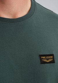 Tamnozeleni pamučni majica s okruglim izrezom, koja ima oznaku od tkanine u crnoj i zlatnoj boji s natpisom "PME LEGEND AMERICAN CLASSIC."