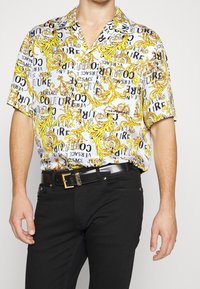 Mies, joka käyttää lyhythihaista nappipaitaa, jossa on keltaisia barokkikuvioita sekä Versace Couture -teksti, yhdistettynä mustiin housuihin ja vyöhön.