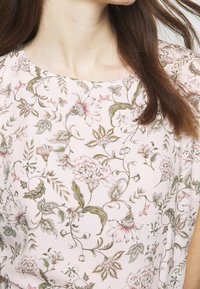 Svetle růžová květinová blůza s jemnými vzory květin a listů v tlumených zelených a měkkých růžových odstínech, s kulatým výstřihem a splývavým materiálem.