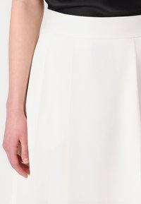 White pleated skirt with a smooth texture featuring a wide waistband and vertical stripes, partially visible with a hand resting on the fabric.