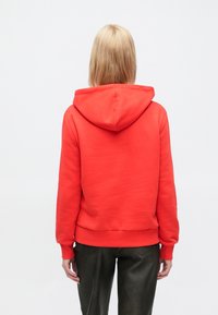 Κόκκινο φούτερ με ευρύχωρη κουκούλα, ριμπ μανσέτες και τελείωμα, κατασκευασμένο από απαλό ύφασμα, εμφανίζεται από πίσω, σε συνδυασμό με σκουρόχρωμο παντελόνι.