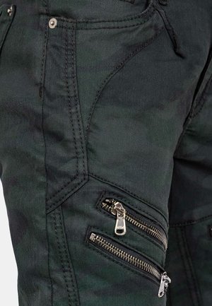 Le pantalon cargo en camouflage noir présente un design ajusté avec plusieurs poches zippées. Le matériau est durable, avec des détails de coutures visibles et des zips.