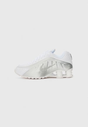 Zapatilla deportiva blanca con acentos de satén plateado, detalles perforados, punta redondeada, diseño con cordones y columnas de amortiguación traseras distintivas.