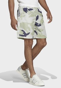 Pantaloni corti mimetici in verde chiaro e blu navy, realizzati in tessuto leggero. Presentano una cintura elastica e strisce laterali. Abbinati a sneakers bianche.