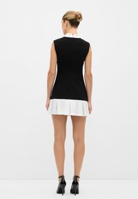 Schwarzes ärmelloses Kleid mit einem weißen plissierten Saum, ausgestattet mit einem hohen Kragen und einem Reißverschluss hinten. Getragen mit schwarzen Absatzschuhen.