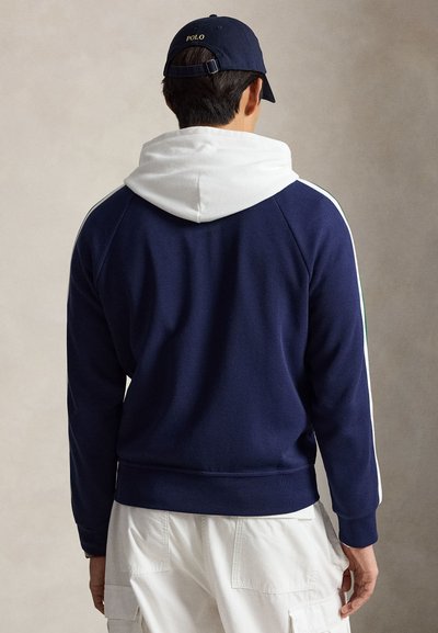 Homme portant un sweat-shirt bleu marine avec capuche blanche et bandes blanches sur les manches, une casquette bleu marine et un pantalon cargo blanc, vu de dos sur un fond uni.