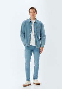 Chaqueta de mezclilla azul claro sobre una camiseta blanca, combinada con jeans azules claros y zapatillas beige, con un bolsillo frontal y cierre de botón.