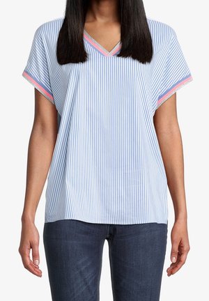 Vrouw met een blouse met korte mouwen in lichtblauw en wit met verticale strepen, en roze- en zilverkleurige randafwerking op de mouwen en V-hals, gecombineerd met een donkere spijkerbroek.