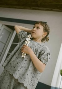 Top in cotone grigio a motivo occhiello con orlo scallop, abbinato a un pantalone coordinato. Bambino che tiene una collana di perline naturali, in un ambiente interno.