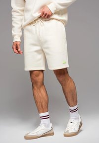 Shorts en polaire couleur crème avec une taille élastique et un cordon de serrage, arborant un petit patch de logo vert. Baskets blanches avec des accents beiges.