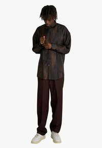 Camicia marrone con bottoni in tessuto strutturato, maniche lunghe e vestibilità rilassata, abbinata a pantaloni bordeaux e sneakers bianche.